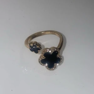 Guldfärgad ring med svarta blommor - Superfin guldfärgad ring med två svarta blommor i olika storlekar. Blommorna har en glittrig kant som ger extra bling. Ringen är öppen framtill och har en modern, trendig design som sticker ut.