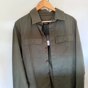 Olivgrön overshirt  - Stilren olivgrön overshirt från Threadbare med klassisk krage, bröstfickor med lock och knäppning framtill. Helt ny