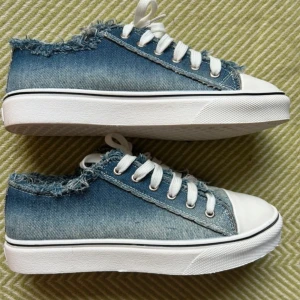 Blå denim sneakers med fransad kant - Snygga sneakers i blått denimtyg med fransade kanter för en cool och avslappnad look. Skorna har vita snören, vit tå och platt vit sula. 