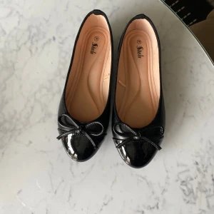 Svarta ballerinaskor med rosett  - Snygga svarta ballerinaskor  med glansig finish och söt rosett framtill. Skorna har rund tå och platt sula, perfekta för en stilren look. Tillverkade i syntetmaterial med mjuk innersula för extra komfort. Använda en gång, så i fint skick 