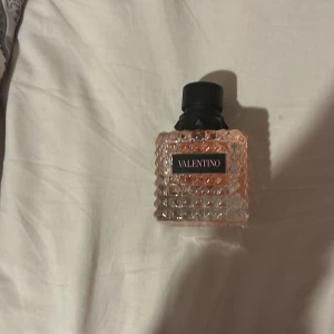 Valentino parfym med nitar - Säljer en lyxig parfym från Valentino i en fyrkantig glasflaska med nitar och rosa-beige tonad vätska. Flaskan har en svart, matt kork med nitar och en etikett med rosa text. Perfekt för dig som gillar exklusiva dofter och snygg design. Alldrig använd 