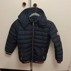 Mörkblå Moncler pufferjacka med huva - Säljer en mörkblå pufferjacka från Moncler med avtagbar huva och coola ränder i rött, vitt och blått på muddar och nederkant. Jackan har dragkedja framtill, två fickor med dragkedja och klassisk Moncler-logga på ärmen. Strl 126 alltså passar de bättre om du är ”petit” eller för barn upp till 12 år