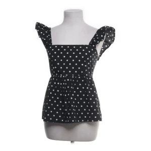 Svart prickig blus med volangärmar - Supersöt svart blus med vita prickar och volangärmar. Toppen har fyrkantig ringning och smock baktill för extra komfort. Perfekt för dig som gillar en lekfull och trendig stil. Luftigt material och lätt utställd passform.