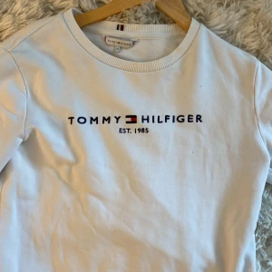 Vit sweatshirt från Tommy Hilfiger  - Vit sweatshirt från Tommy Hilfiger i bomull med broderad logga och text på bröstet. Klassisk rund halsringning, ribbade muddar och nederkant samt diskreta Hilfiger-detaljer vid ärmslut och insida. Perfekt för en clean och stilren look.