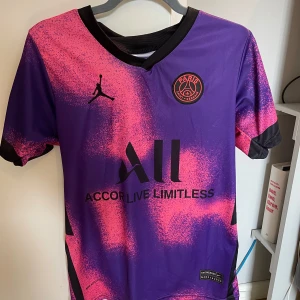 PSG Messi #30 lila/rosa fotbollströja - Säljer en PSG fotbollströja med Messi #30 på ryggen. Tröjan är lila med rosa detaljer i ett spraymönster, svarta muddar och loggor. Officiell Jordan x PSG collab, tillverkad i lätt och ventilerande material. Perfekt för dig som älskar fotboll och Messi.