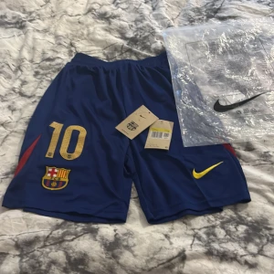 Blå FC Barcelona shorts Nike S - Snygga blå FC Barcelona shorts från Nike med nummer 10 och klubbmärke framtill. Gula och röda detaljer, elastisk midja med vit snörning. Perfekta för fotbollsträning eller att bara chilla i. Lätta och sköna i polyester. Allt är nyskick förutom att det är ett litet slitage högst upp vid stängningen av påsen (märks inte av).