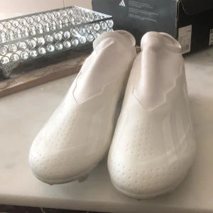 Vita adidas X Crazyfast fotbollsskor - Säljer ett par helvita adidas X Crazyfast fotbollsskor med strumpliknande skaft och diskret adidas-logga på sidan. Skorna har en slimmad design, plös utan snörning och greppig sula med fasta dobbar för spel på gräs. Perfekta för dig som vill sticka ut på planen. De har bara använts en gång och dem är som om dem var helt nya fortfarande.
