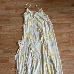 Lång blommig volangklänning i pastell - Superfin långklänning med tunna axelband och volangdetaljer. Klänningen har ett blommigt mönster i ljusa pastellfärger som gult, lila och vitt. Perfekt flowy modell med romantisk känsla och asymmetriska volanger längs hela klänningen.