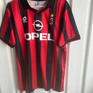 AC Milan Baggio 18 Lotto fotbollströja - Klassisk AC Milan fotbollströja med Baggio och nummer 18 på ryggen. Röd och svart med breda vertikala ränder, broderat klubbmärke och Opel-sponsor på bröstet. Kortärmad modell med svart krage och detaljer. Perfekt för dig som älskar retro och italiensk fotboll.