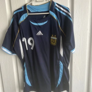 Argentina bortatröja Adidas VM 2006 - Säljer en officiell Argentina bortatröja från VM 2006 med nummer 19. Tröjan är mörkblå med ljusblå och vita detaljer, guldiga linjer och broderat förbundsmärke. Adidas-logga på bröstet och officiellt VM-märke på ärmen. Tillverkad i lätt och ventilerande material.