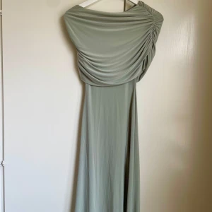 Ljusgrön långklänning med drapering - Snygg ljusgrön långklänning med en axel och draperad överdel. Klänningen har en elegant, flowy siluett och är tillverkad i ett mjukt, slätt material som faller fint. Perfekt för dig som vill sticka ut med en stilren och modern look. Oanvänd 