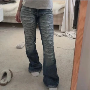 Blå bootcut jeans med slitningar - Säljer ett par blå bootcut jeans med kraftiga slitningar framtill. Jeansen har en klassisk femficksmodell och är tillverkade i denim. Perfekta för dig som gillar en edgy och trendig stil.
