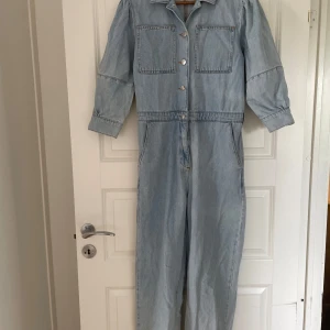 Ljusblå jeansjumpsuit från NA-KD - Säljer en snygg ljusblå jeansjumpsuit från NA-KD i storlek 36. Jumpsuiten har stora bröstfickor, knappar framtill, krage och långa ärmar med knappdetaljer. Tillverkad i klassiskt denimtyg och har bakfickor för extra stilpoäng.