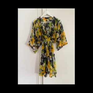 Blommig kortklänning med knytband - Superfin kortklänning / tunika med stora gula och gröna blommor på svart och vit botten. Klänningen har v-ringning, vida trekvart ärmar och knytband i midjan för en snygg passform. Perfekt för sommarens varma dagar och ger en riktigt fräsch vibe. Snygg till vardags som fest och till stranden!