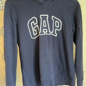 Mörkblå hoodie från GAP - Mörkblå hoodie från GAP med stor vit broderad logga på bröstet. Klassisk ficka framtill och huva. Mjuk bomullsblandning och långärmad modell, perfekt för chill dagar eller när du vill ha en enkel och snygg look.