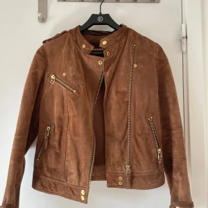 Brun mockajacka från Mango - Snygg brun skinnjacka i mocka från Mango med guldiga dragkedjor och knappar. Jackan har flera fickor med dragkedja och axelremmar. Nypris var 599 kr och är oanvänd utan prislapp💕