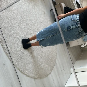 Blå jeanskjol med slits - Säljer en snygg blå lång jeanskjol med slitningar och en hög slits framtill. Kjolen har klassiska fickor, bälteshällor och råa kanter nertill för en cool vibe. Perfekt för dig som gillar streetstyle och vill sticka ut lite extra.