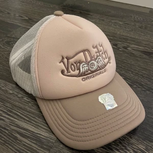 Beige Von Dutch truckerkeps - Snygg truckerkeps från Von Dutch i beige och ljusrosa med broderad logga framtill. Kepsen har vit mesh baktill för extra ventilation och justerbar snapback. Perfekt accessoar för en cool och avslappnad stil.