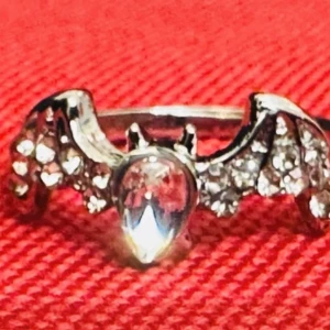 Ny silvrig ring med fladdermus av strass & kristaller – gotisk statement -  Detaljrik ring i silvrig metall med glittrande kristaller och fladdermusvingar. Den droppformade stenen i mitten ger ringen en dramatisk och alternativ känsla. Perfekt för dig som gillar gotisk estetik eller halloweeninspirerade accessoarer. Innerdiameter: ca 2,1 cm (EU 62). Ny utan prislapp. Priset är fast – jag prutar inte, men erbjuder rabatt vid köp av flera saker. Väl värt att kika på mina andra annonser!  ⸻  