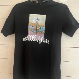 Svart t-shirt H&M Restless Youth XS - Svart t-shirt från H&M med avslappnad passform och tryck framtill med texten 'Restless Youth' och en cool graffitibild. Klassisk rund hals och korta ärmar. Perfekt för dig som gillar streetstyle och vill sticka ut lite extra.