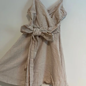 Beige omlottklänning med rosett - OBS! Väldig dåliga bilder kan skicka hur den sitter på. Beige kortklänning i linne med omlottmodell och stor knytrosett i midjan. Klänningen har smala axelband som är justerbara och v-ringad urringning, perfekt för varma dagar. Lätt och luftig känsla med dragkedja i ryggen.