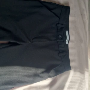 Svarta kostymbyxor från 157 - Klassiska svarta kostymbyxor från 157 med raka ben och bälteshällor. Byxorna har en enkel knappstängning och diskreta sidfickor. Perfekta för en clean och stilren look. Materialet känns mjukt och följsamt.