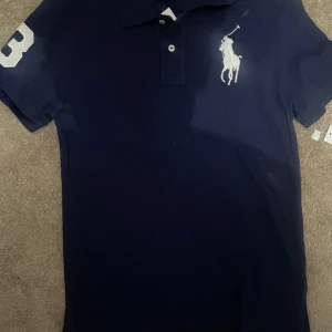 Marinblå pikétröja Polo Ralph Lauren - Snygg marinblå pikétröja från Polo Ralph Lauren med klassisk krage, två knappar och stor vit logga på bröstet. På ena ärmen finns en vit siffra 3. Tröjan är gjord i bomull och har korta ärmar. Perfekt för en clean och sportig look.