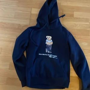 Mörkblå Polo Bear hoodie från Ralph Lauren - Snygg mörkblå hoodie från Polo Ralph Lauren med ikoniska Polo Bear-trycket på bröstet. Tröjan är använd ett fåtal gånger. Tröjan har huva med snörning och långa ärmar. Perfekt för dig som gillar streetstyle och vill ha något klassiskt men ändå lekfullt.