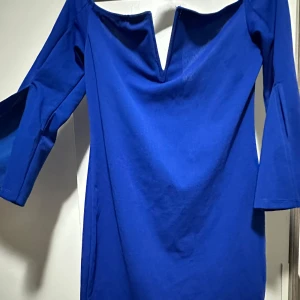 Kornblå offshoulder kortklänning - Kornblå kortklänning med offshoulder och djup v-ringning framtill. Klänningen har trekvartsärmar och är sydd i ett mjukt, slätt tyg som känns bekvämt mot huden. Enkel och stilren design som sticker ut med sin starka färg.