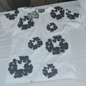 Vit t-shirt med svart blommigt tryck Denim Tears - Snygg vit t-shirt från Denim Tears med unikt svart blommigt tryck över hela plagget. Klassisk passform med rund hals och korta ärmar. Tillverkad i mjuk bomull som känns skön mot huden. Perfekt för dig som vill sticka ut med en cool och modern look.