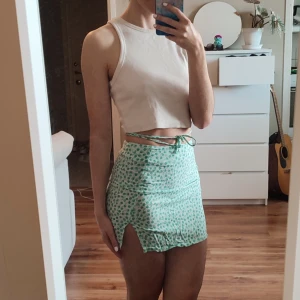 Mintgrön blommig kjol - Supersöt mintgrön kortkjol från FB Sister i storlek XS. Kjolen har grönt blommönster, slits på ena sidan och knytband i midjan som ger en extra snygg detalj. Perfekt för vår och sommar när du vill sticka ut lite extra!
