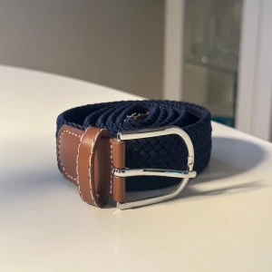Mörkblå flätat bälte 100cm - Snyggt mörkblått flätat bälte med bruna skinn-detaljer och silvrigt spänne. Bältet har en klassisk look med vit kontrastsöm på skinnet och passar perfekt till jeans eller chinos. Flexibelt material som gör att den kan justeras enklare. Bältet är 100cm långt