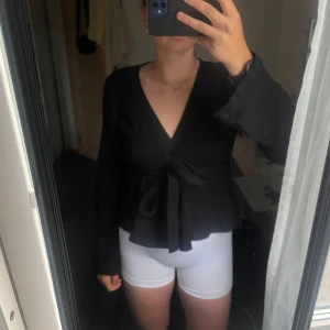Svart omlottblus med knytband - Säljer en svart omlottblus med v-ringning och långa, vida ärmar. Blusen har ett knytband i midjan som ger en snygg siluett och en flowy känsla. Perfekt att styla med jeans eller kjol för en trendig look.
