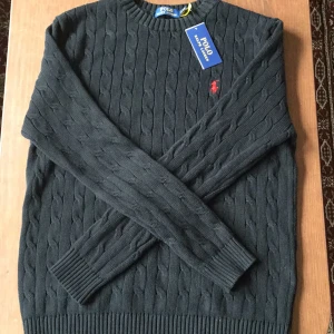 Svart kabelstickad tröja Polo Ralph Lauren - Kabelstickad svart tröja från Polo Ralph Lauren med klassisk rund hals och röd broderad logga på bröstet. Tröjan har ribbade muddar och är tillverkad i mjuk bomull. Perfekt för dig som gillar stilren och tidlös design.