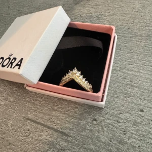 Guldfärgad ring med stenar från Pandora - Snygg guldfärgad ring från Pandora med V-formad design och glittrande stenar längs hela ovansidan. Ringen har en elegant och modern look med flera små vita stenar som ger extra bling. Perfekt för dig som vill sticka ut med dina accessoarer. (Helt oanvänd och perfekt för dig som tycker om accessoarer) 