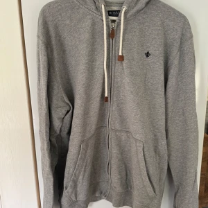 Grå hoodie från Morris med dragkedja - Snygg grå hoodie från Morris med dragkedja och huva. Tröjan har vita snören med bruna detaljer, två fickor fram och en liten broderad logga på bröstet. Priset kan diskuteras vid snabb och smidig affär. 