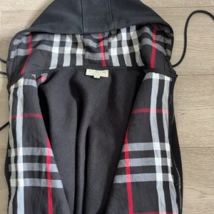 Svart Burberry kofta med rutig lining - Svart kofta från Burberry med klassiskt rutigt foder i svart, vitt och rött. Jackan har huva med dragsko, dragkedja framtill och en ficka. Materialet är mjukt och känns som bomull eller polyester. Perfekt för dig som vill ha en stilren jacka med ikonisk Burberry-touch. Dragkedjan är ganska dålig därför jag säljer den lätt att byta ut dock kostar max 30 kr har bara inte haft tid med det