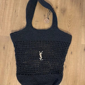 Svart stickad shoppingväska YSL - Snygg svart stickad shoppingväska från YSL med klassisk logga i silver framtill. Väskan har ett öppet, nätliknande parti i mitten och breda handtag som gör den enkel att bära på axeln. Perfekt för dig som vill ha en trendig och rymlig väska.