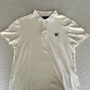 Vit pikétröja från Lyle & Scott  - Klassisk vit pikétröja från Lyle & Scott i storlek M. Tröjan har korta ärmar, krage med tre knappar och den ikoniska gula fågel-loggan broderad på bröstet. Tillverkad i mjuk bomull som känns skön mot huden. Perfekt för en clean och stilren look.