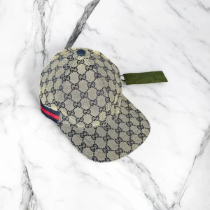 Gucci keps med GG-mönster och rödblå rand - Exklusiv keps från Gucci i beige och marinblått med klassiskt GG-monogram över hela kepsen. Snygg röd och marinblå rand på sidan. Justerbar rem baktill och böjd skärm. Tillverkad i bomullscanvas med innerfoder i marinblått tyg. Perfekt för dig som gillar lyxiga accessoarer.