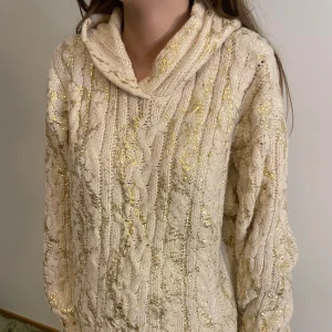 Beige stickad hoodie med guldtråd - Supermysig beige stickad hoodie med flätmönster och inslag av glittrig guldtråd. Tröjan har en huva och långa ärmar, perfekt för dig som gillar en chill och snygg look med lite extra bling.