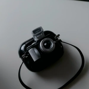 Airport pro cover i kameradesign - Unik svart axelväska formad som en retro kamera med silverdetaljer och coola knappar. Väskan har ett hårt skal och ett justerbart axelband. Perfekt för dig som vill sticka ut och gillar fotostil. Rymmer småsaker som hörlurar eller nycklar.