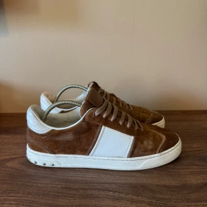 Valentino Rockstud sneakers mocka brun/vit - Snygga Valentino Rockstud sneakers i brun mocka med vita skinnpaneler och klassiska nitar på sulan. Låga med rund tå, platt sula och snörning. Perfekta för dig som vill ha en lyxig och stilren look med streetkänsla.