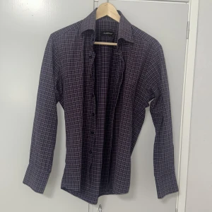 Lila rutig skjorta från Ruben's M - Snygg lila skjorta från Ruben's i storlek M med rutigt mönster i mörka toner. Skjortan har klassisk krage, långa ärmar och knäppning framtill. Perfekt för dig som gillar stilrena och tidlösa plagg med lite edge.