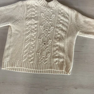 Beige stickad tröja från 157 - Beige stickad tröja från 157 med grovt flätmönster framtill och strukturstickade sidor. Tröjan har hög ribbad krage och långa ärmar. Perfekt för lager-på-lager och chill höst- eller vinterstil.