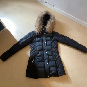 Svart Rockandblue dunjacka med fuskpäls - Används inte ny pris kan även diskuteras snygg svart dunjacka från Rockandblue med quiltade detaljer och lång passform. Jackan har huva med fuskpäls, dragkedja och knappar framtill samt flera fickor med dragkedja. Perfekt för kalla dagar och riktigt stilren look.