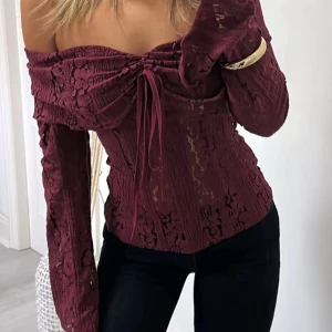 Vinröd offshoulder spetsblus - Vinröd blus i spets med offshoulder och långa ärmar. Toppen har snörning framtill som ger en snygg detalj och figurnära passform. Perfekt för dig som vill sticka ut med en romantisk och trendig look.