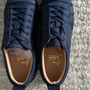 Mörkblå sneakers från Christian Louboutin - Snygga mörkblå sneakers från Christian Louboutin i mocka med klassisk röd sula och rund tå. Skorna har snörning och beige innersula med guldfärgad logga. Perfekta för dig som vill ha en exklusiv och stilren look.