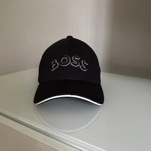 Svart keps från BOSS med vit detalj - Svart keps från BOSS med broderad logga framtill och justerbart spänne bak i metall. Kepsen har en böjd skärm med vit kant och är tillverkad i ett ventilerande polyestertyg. Perfekt för en clean och sportig look.