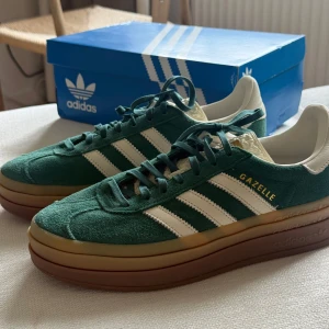Adidas Gazelle grön mocka sneakers - Adidas Gazelle sneakers i mörkgrön mocka med vita detaljer och klassiska tre ränder på sidorna. Platt gummisula i brunt med coolt mönster undertill. Skorna har snörning och guldfärgad Gazelle-text på sidan. Perfekt för dig som gillar retrovibbar och streetstyle. Skorna är oanvända. 